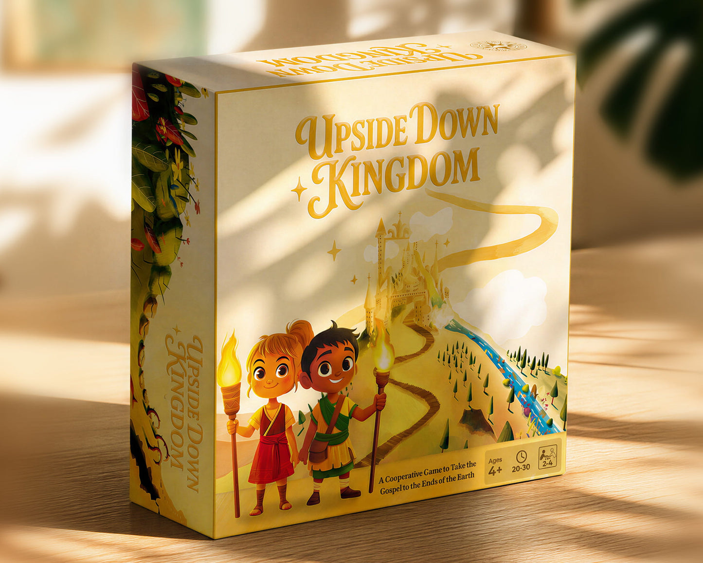 The board game box for 'Upside Down Kingdom' is shown standing on a wooden surface with warm sunlight and leaf shadows. The box features a cream-colored background with gold lettering and colorful illustrations of two children holding torches, a castle, and a winding path.